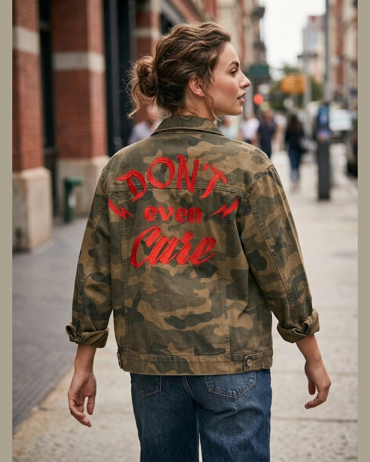 A woman viewed from behind wearing a camouflage jacket with 'DON'T even Care' embroidered in red across the back, standing on a city street.