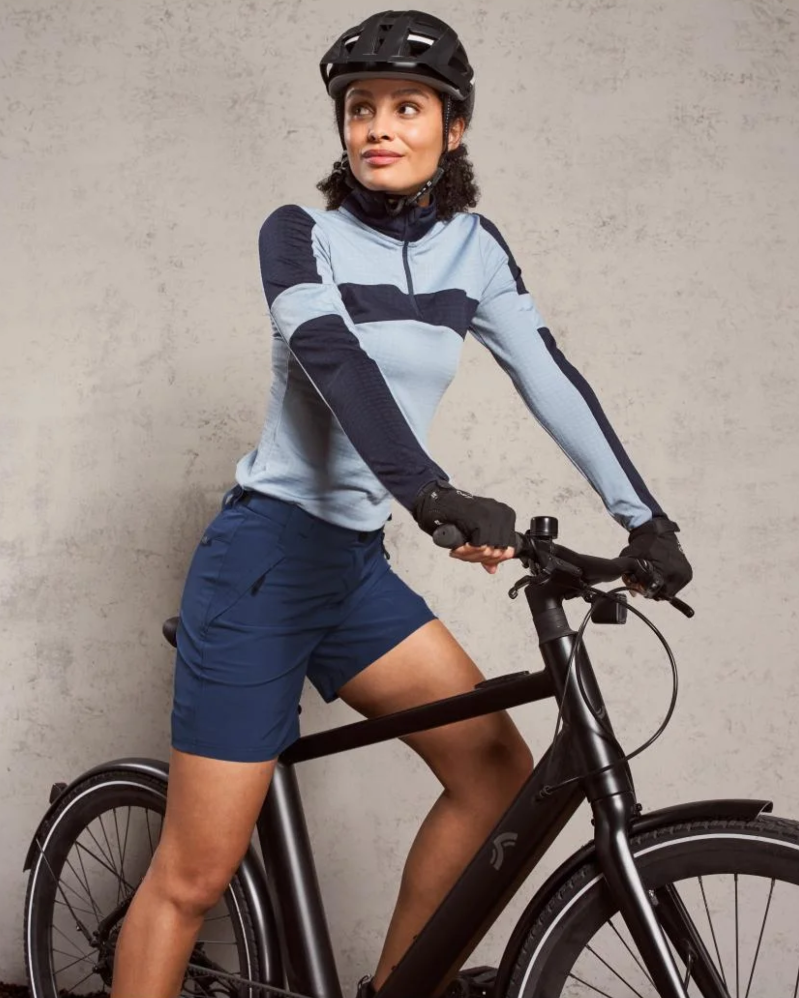 Woman on a bicycle wearing a light blue and navy long-sleeve top and dark shorts against a gray background