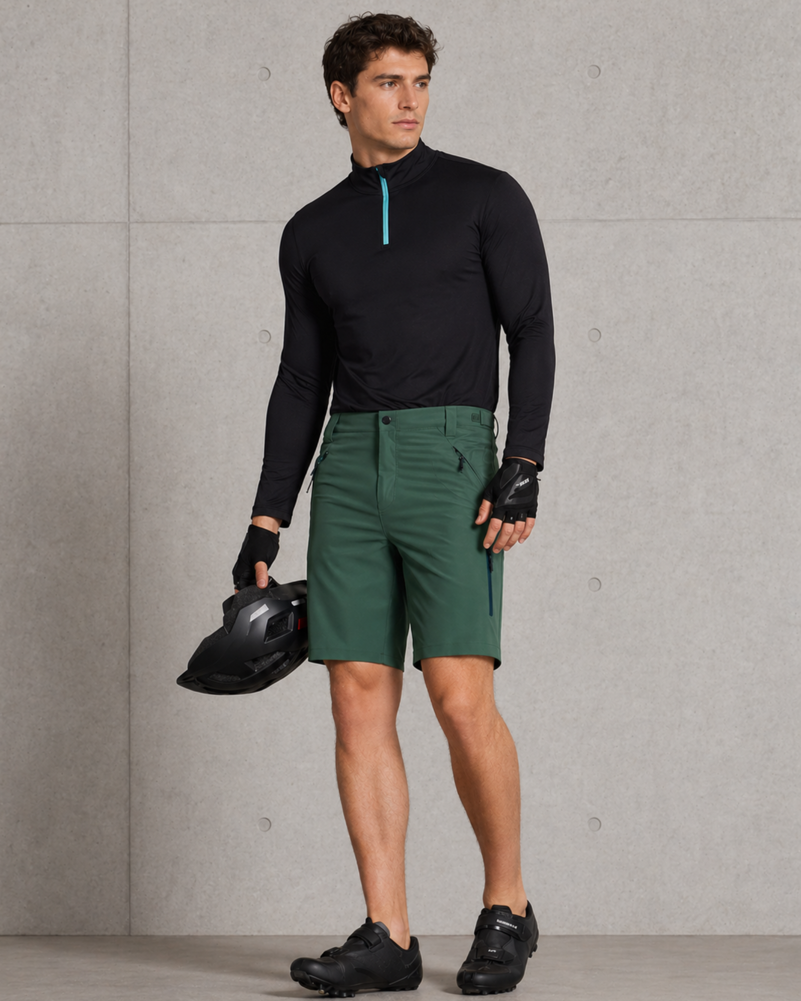Man wearing a black long-sleeve top and green shorts, holding a helmet against a gray concrete wall.