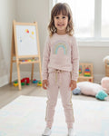 Child wearing a pink outfit with a rainbow design in a room with toys and a whiteboard.