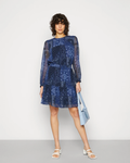 A model stands against a light gray studio background wearing a blue patterned long-sleeve dress with a tie waist and holding a light blue tote bag.