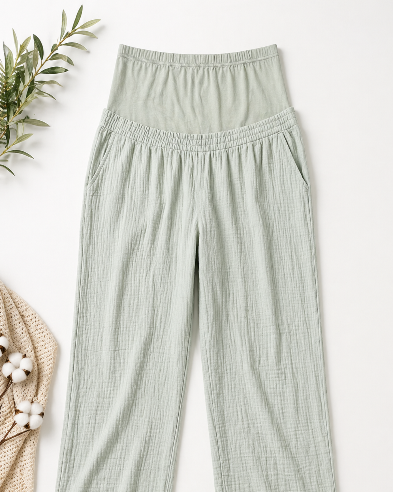 Light green pants on a white background with a plant and woven bag.