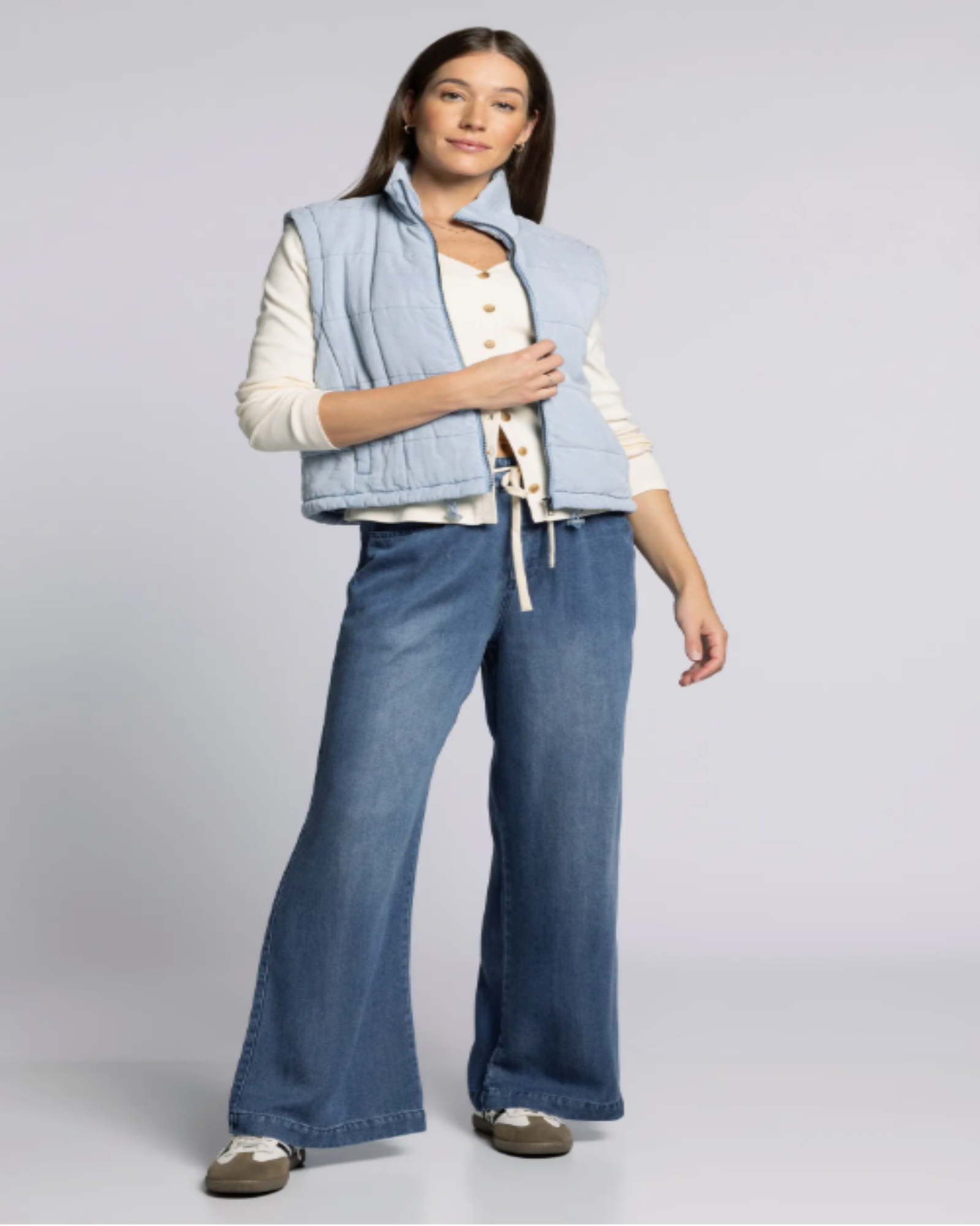 Woman wearing a light blue vest over a white shirt with blue jeans on a gray background