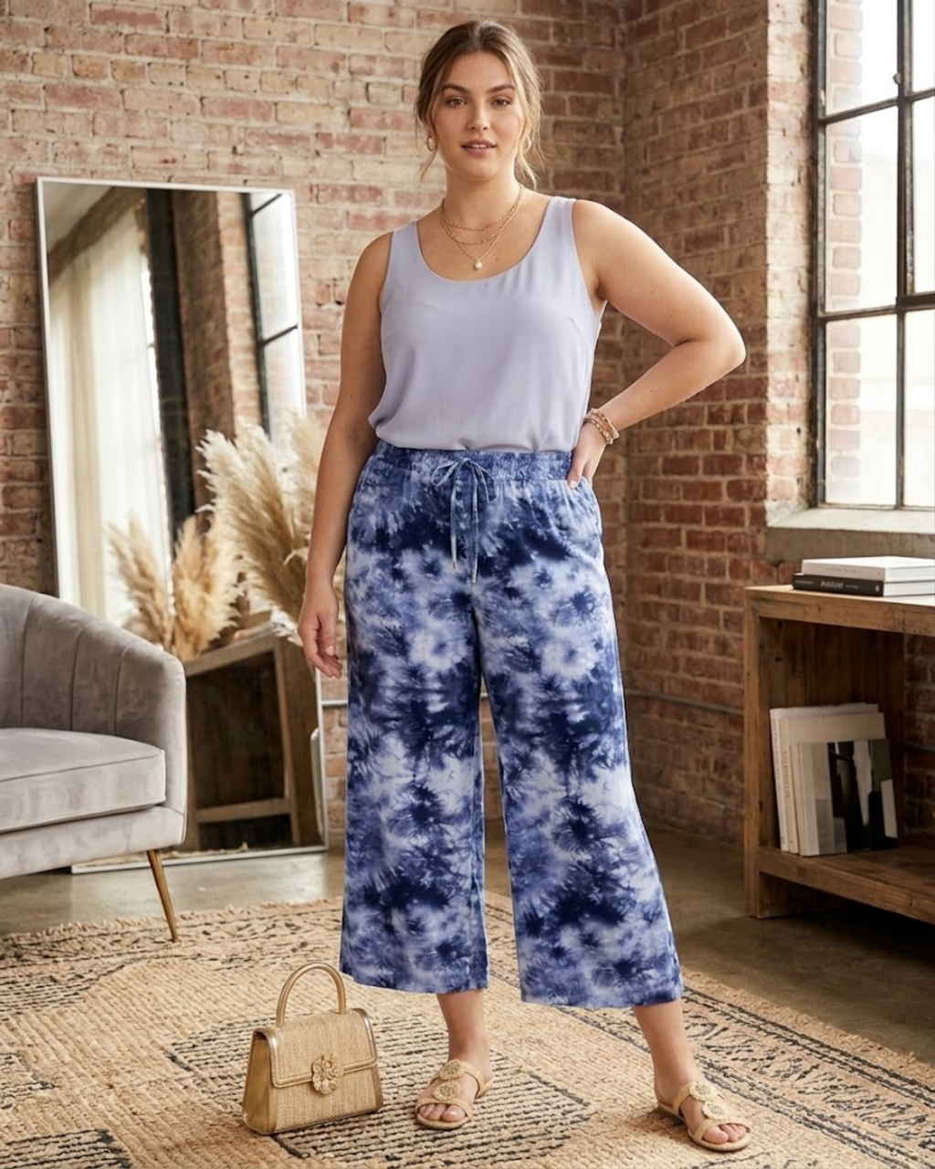 Woman wearing a light blue sleeveless top and tie-dye pants in a room with brick walls and a couch.