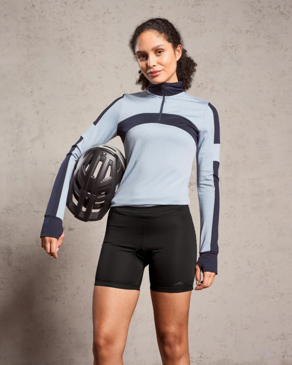 Woman wearing a light blue and black cycling outfit holding a helmet against a gray background