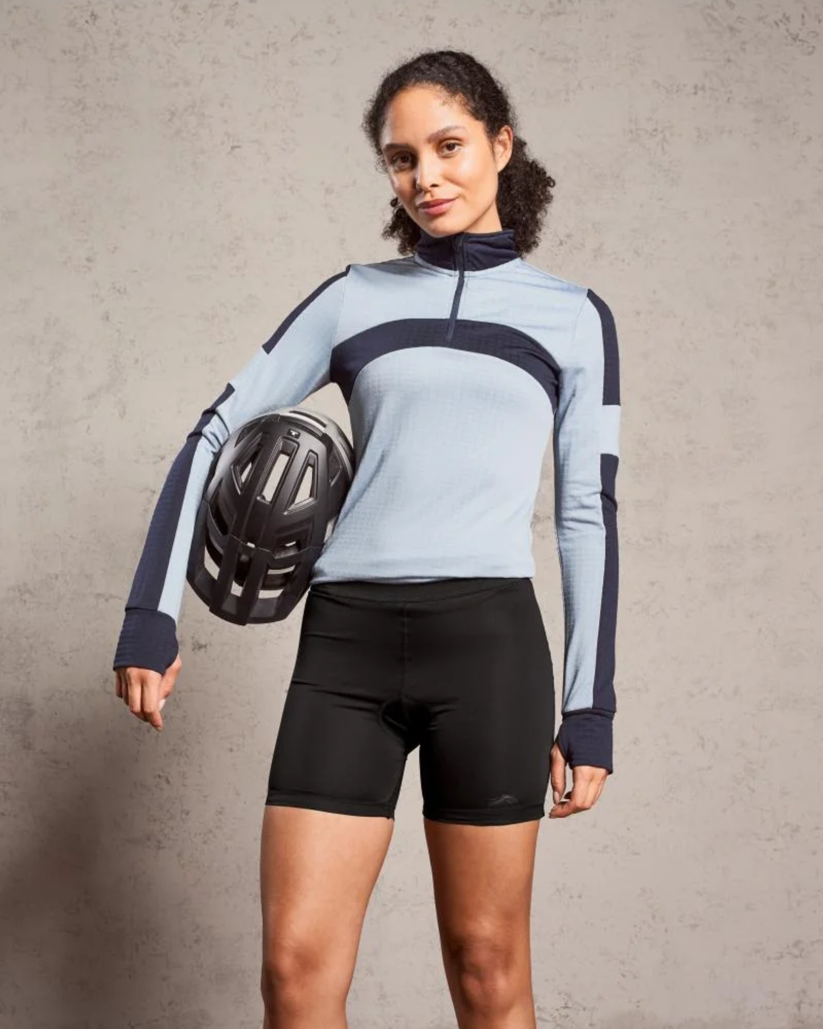 Woman wearing a light blue and black cycling outfit holding a helmet against a gray background